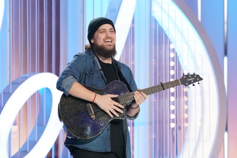 A young man (Oliver steele) playing a guitar and singing on a stage with a microphone stand in front of him. He wears a black shirt and maroon pants while playing guitar.